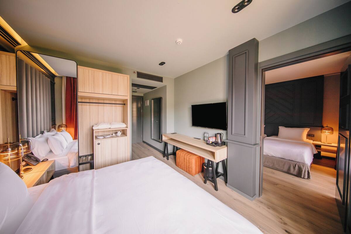 A modern hotel room with a large bed, a desk with a flat-screen TV, a minibar, and a separate sleeping area.
