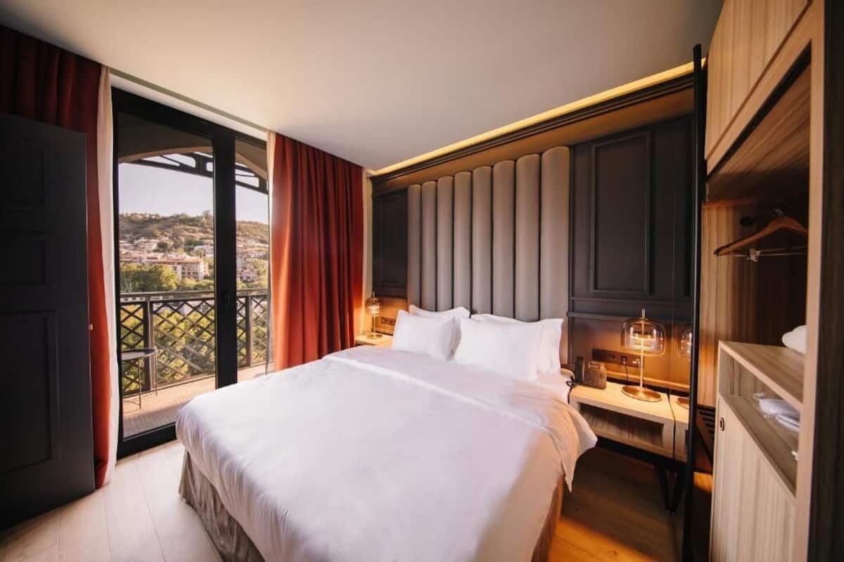 A hotel room with a large bed, a nightstand with a lamp, a wardrobe, and a balcony with a view of the city.