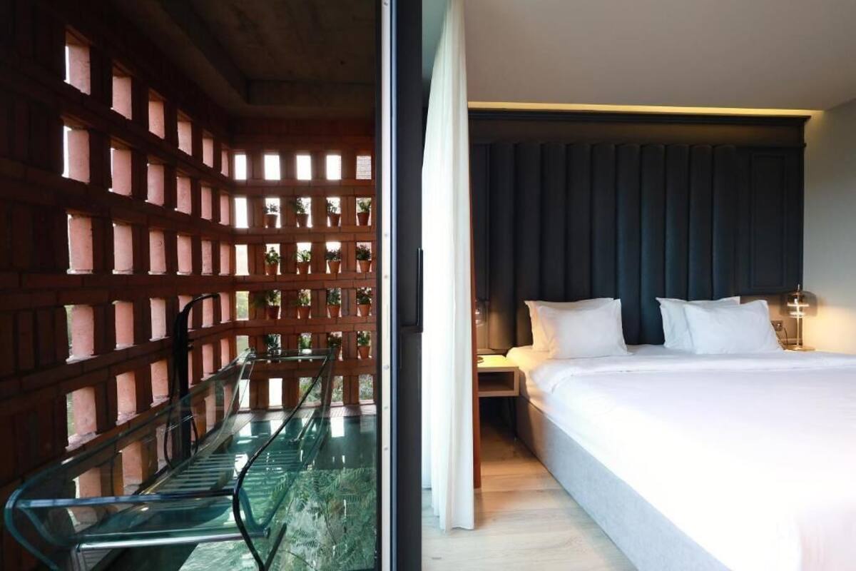 A hotel room with a bed, a glass partition, and a view of a wooden structure with plants.