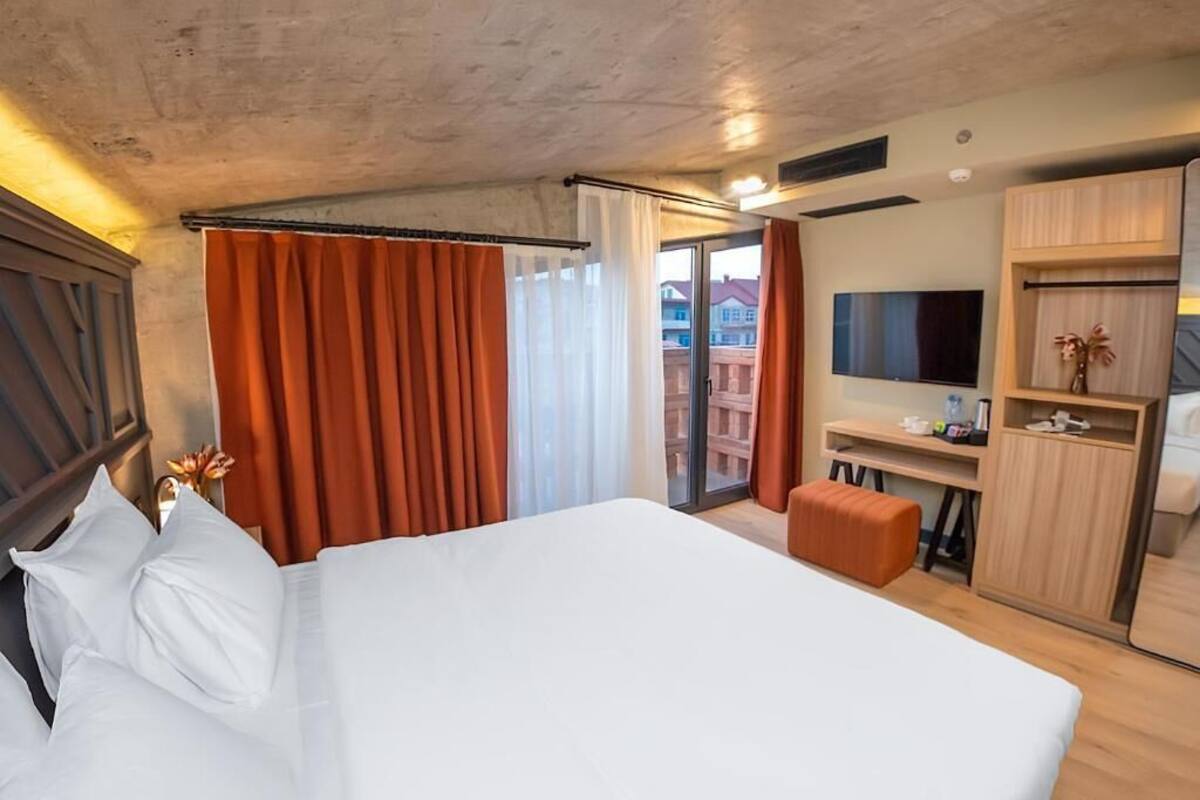 A hotel room with a large bed, a desk with a TV, and a view of buildings through the window.