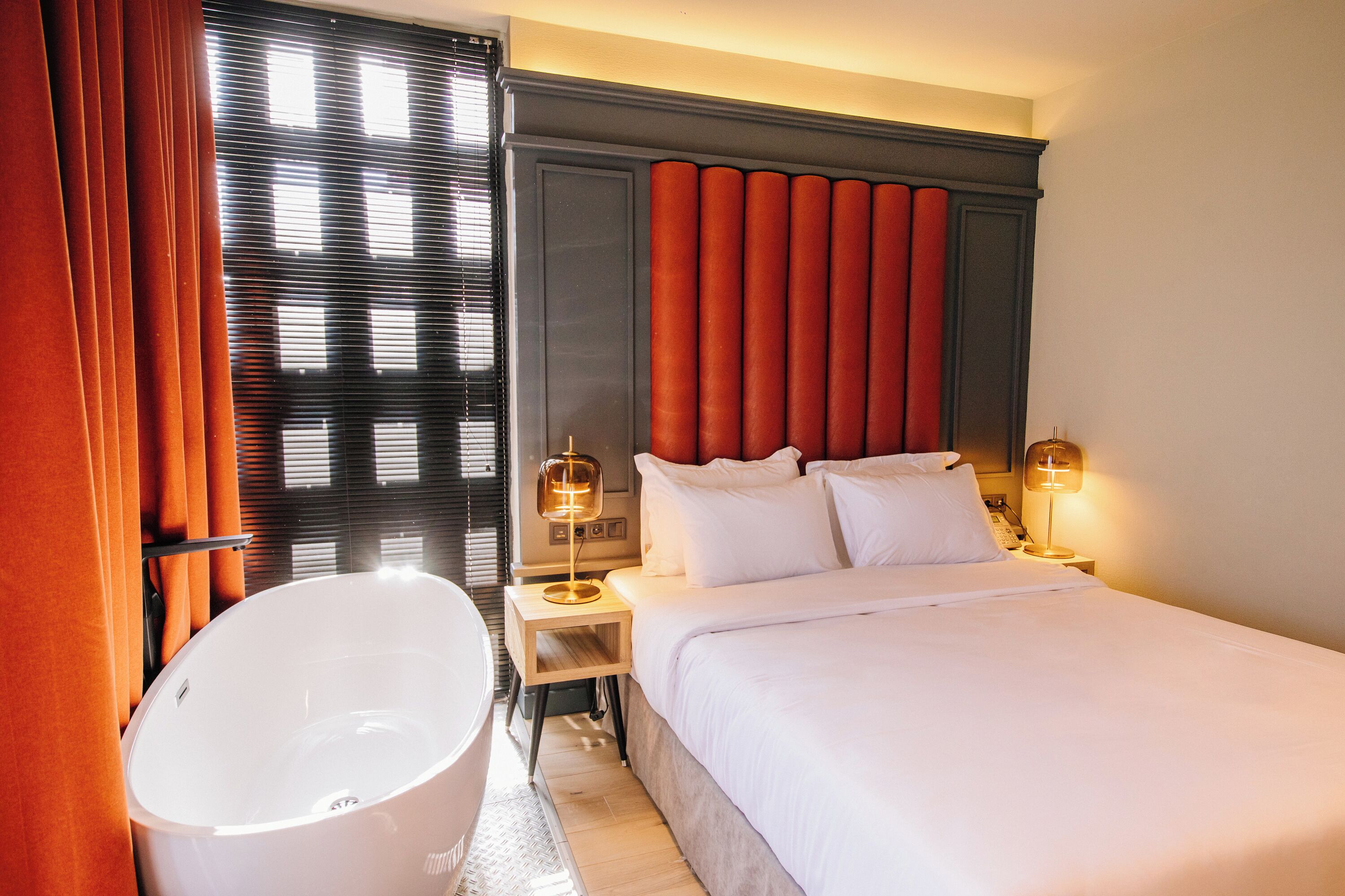 Habitación de hotel con una bañera blanca grande, una cama con sábanas blancas y un cabecero con paneles rojos verticales. Hay dos lámparas de noche en las mesitas.