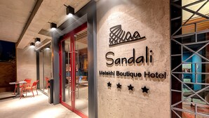 Entrada de un hotel con diseño moderno, que incluye una puerta de cristal, sillas rojas en el exterior y un cartel que dice 'Sandali Metokhi Boutique Hotel'.