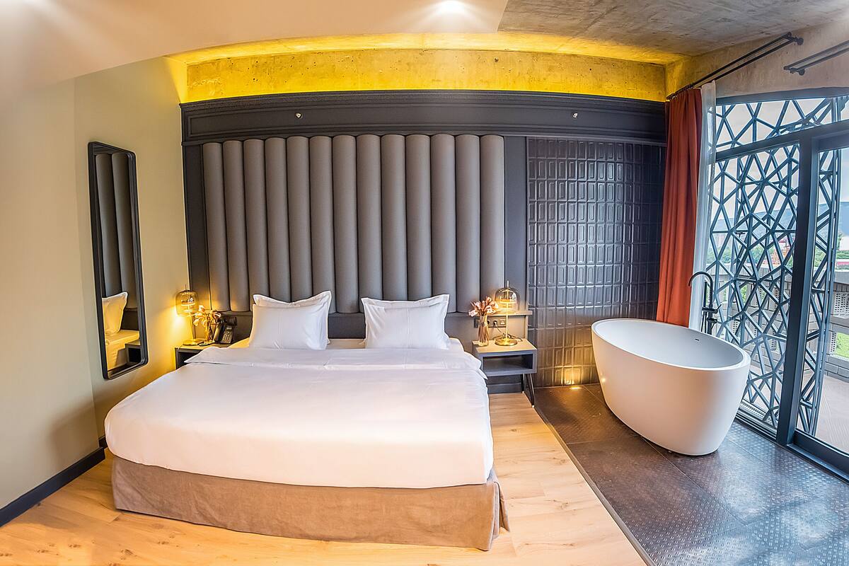 A modern hotel room with a large bed, a bathtub, and a view of the outdoors.