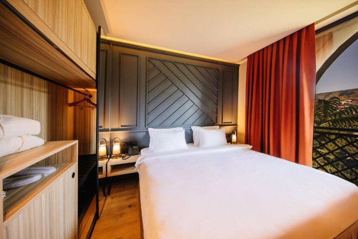 A hotel room with a large bed, a wooden wardrobe, a nightstand with a lamp, and a view of a cityscape through a window with red curtains.