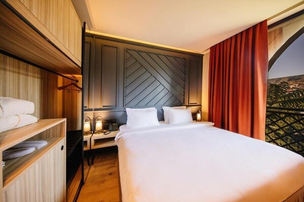 A hotel room with a large bed, a wooden wardrobe, a nightstand with a lamp, and a view of a cityscape through a window with red curtains.
