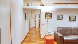 Un couloir au plancher de bois mène à un salon où se trouvent un canapé en cuir marron, un petit pouf orange et une lampe sur pied. Des tableaux encadrés ornent le mur, et une petite table avec une lampe y est également présente.
