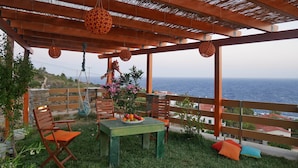 An outdoor seating area with a table and chairs, a hanging swing, and a view of the sea. - Anemi Apartments (Ikaria)