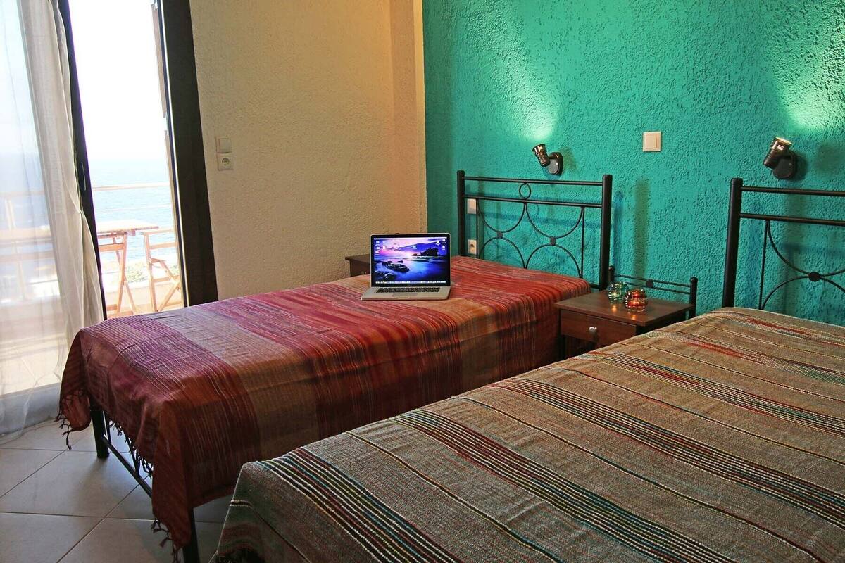 A room with two beds, a laptop on one bed, and a view of the sea through a large window.