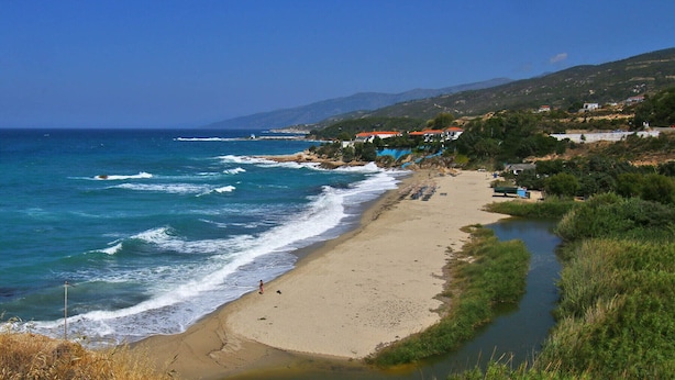 A coastal beach with a sandy shore, a small water channel, and buildings with red roofs in the background. - Anemi Apartments (Ikaria)