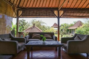 A covered outdoor seating area with a sofa and armchairs, a table with a plant, and a view of traditional buildings and greenery. - Bali Eco Living Yoga Healing Meditation and Retreat Center (Denpasar)