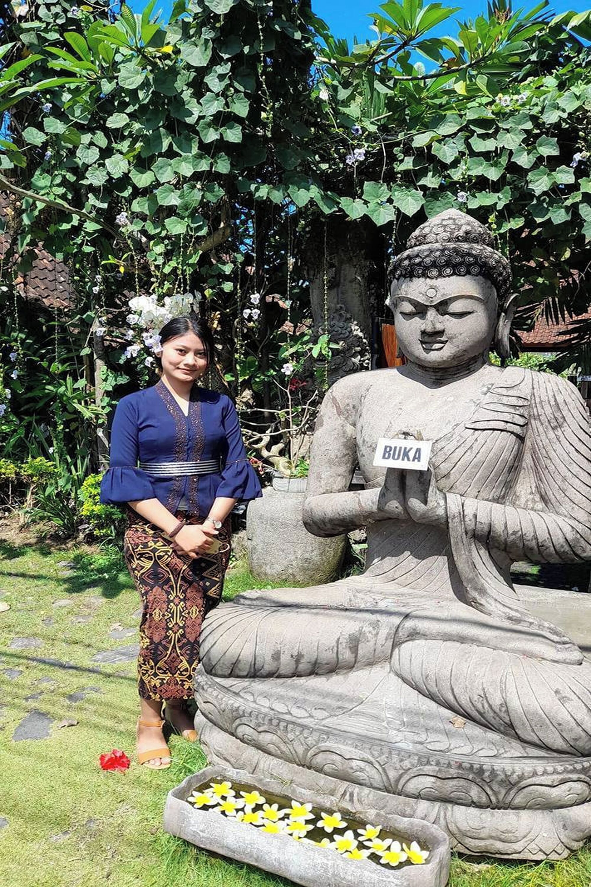 A large stone Buddha statue with a placard reading 'BUKA' in a garden setting.