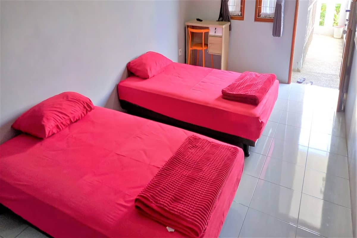 Ein Zimmer mit zwei Betten, jeweils mit roter Bettdecke und Kissen. Es gibt einen kleinen Schreibtisch mit Stuhl und ein Regal.