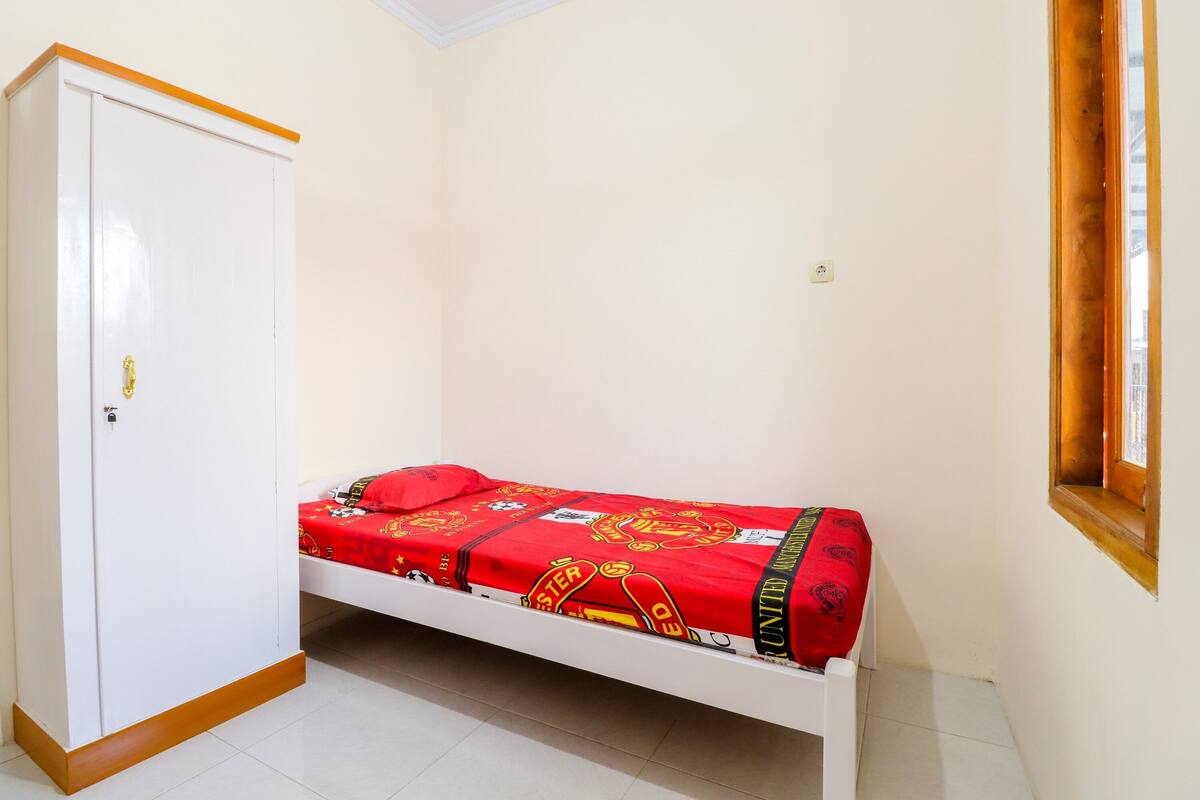 A single-bed room with a white wardrobe, a red bedspread, and a window with a wooden frame.