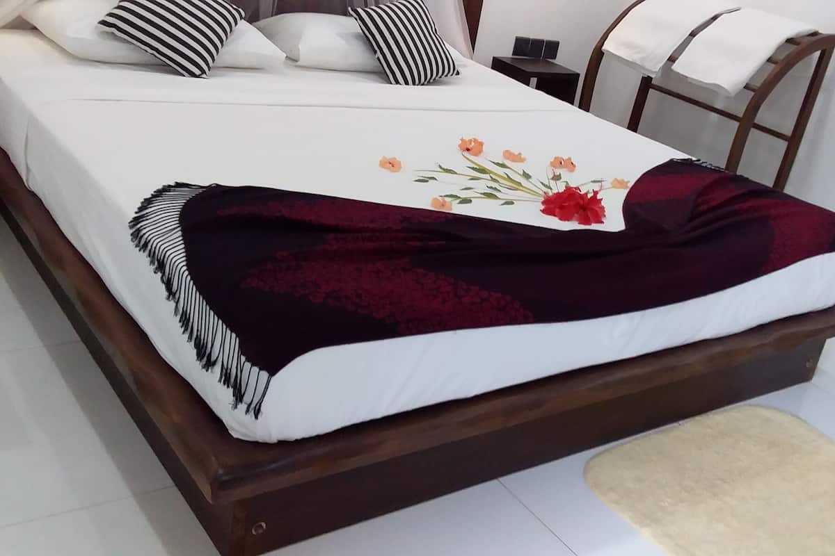 Cama com colchão branco, manta vermelha escura e design floral. Tem cabeceira de madeira, mesa de cabeceira pequena com abajur, e cadeira com toalha branca sobre ela.