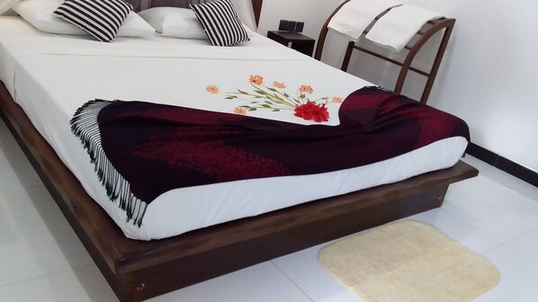 Una cama con colchón blanco, manta rojo oscuro y diseño floral. Se observa un cabecero de madera, una mesita de noche con una lámpara y una silla con una toalla blanca sobre ella.