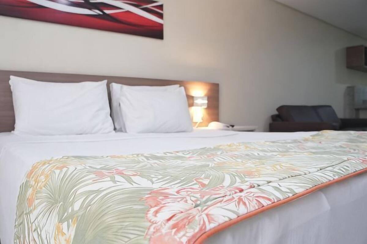A neatly made bed with white linens, a floral-patterned blanket, and a headboard with a light fixture.