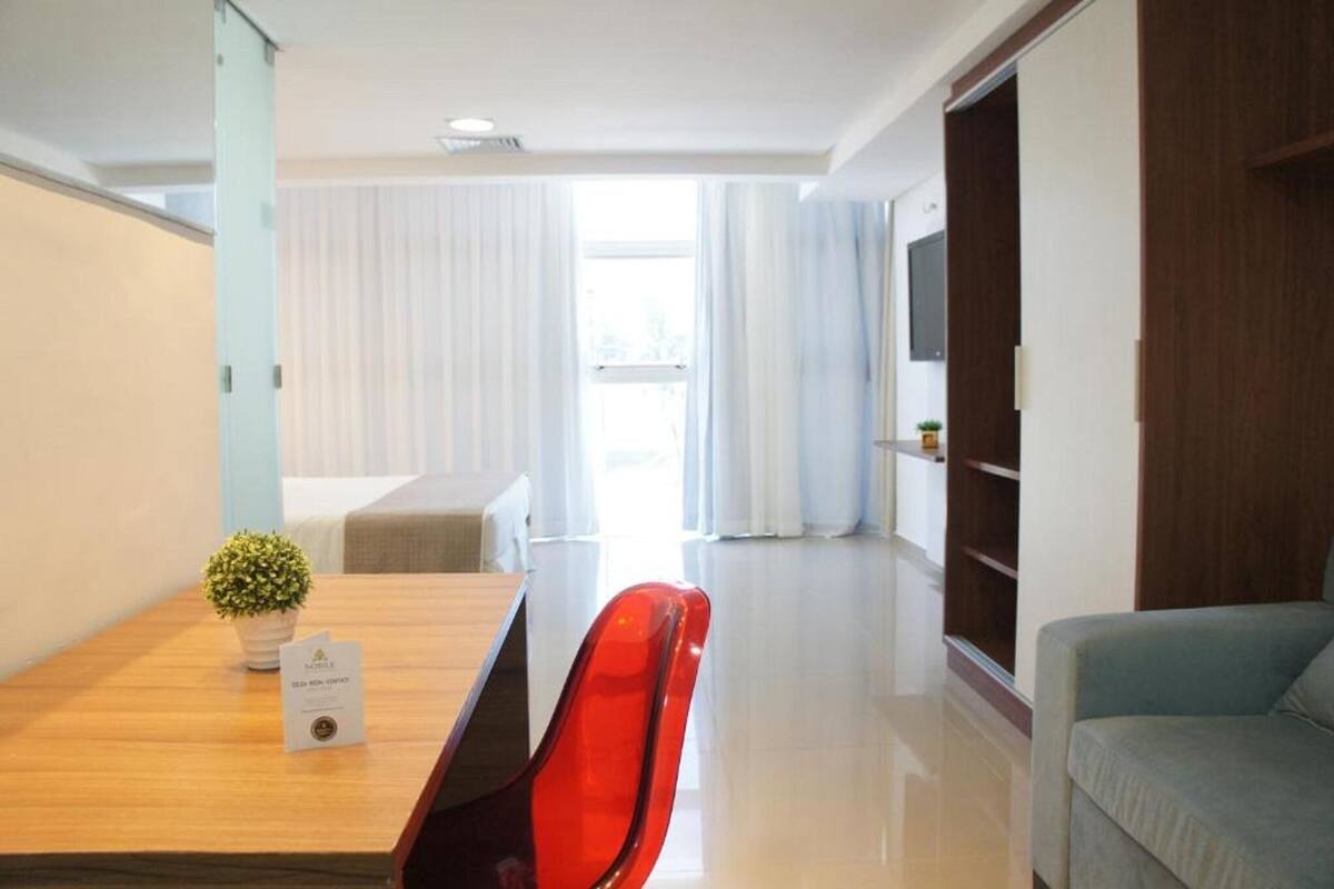 A hotel room with a wooden dining table, a red chair, a grey sofa, a wooden wardrobe, and a potted plant.