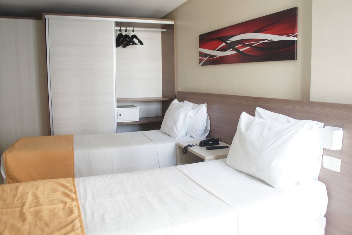 A hotel room with two beds, a wardrobe, a painting, and a telephone.