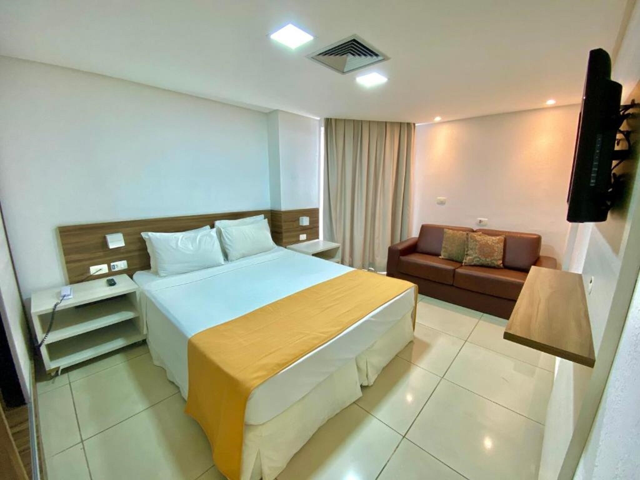 A hotel room with a bed, bedside table, a sofa, a TV mounted on the wall, and a ceiling vent.