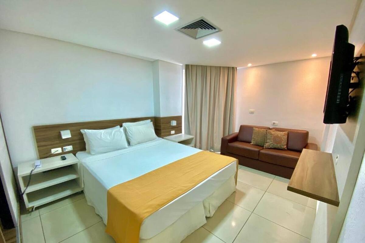 A hotel room with a bed, bedside table, a sofa, a TV mounted on the wall, and a ceiling vent.