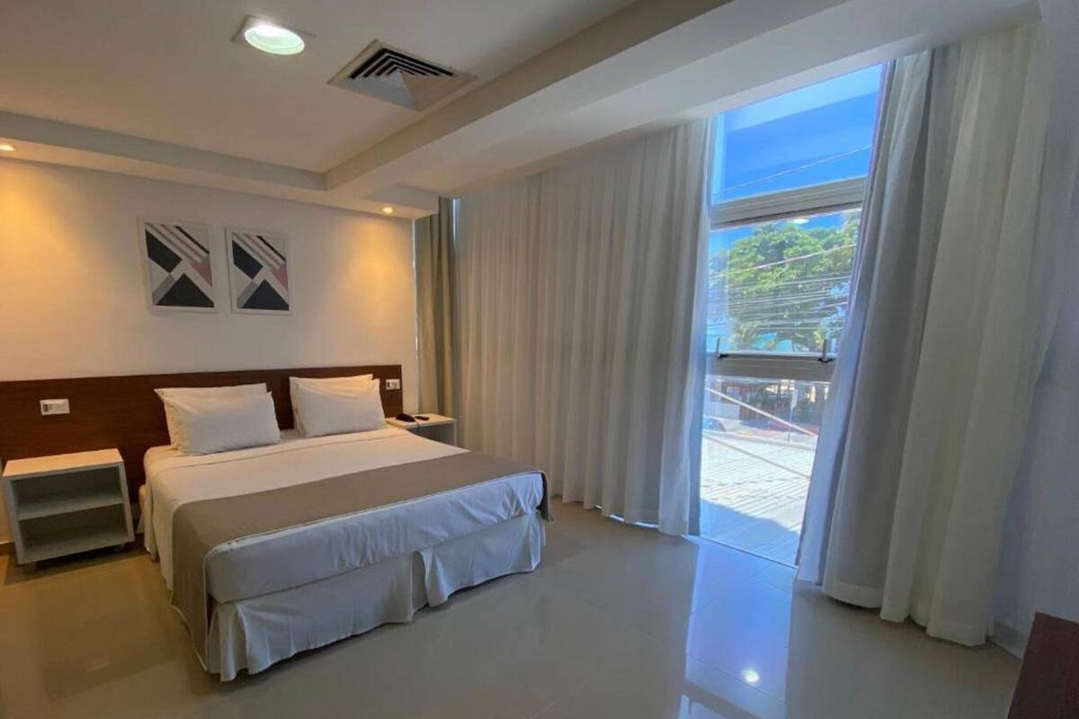 A hotel room with a large bed, a nightstand, a ceiling vent, and a view of the outdoors through a large window.