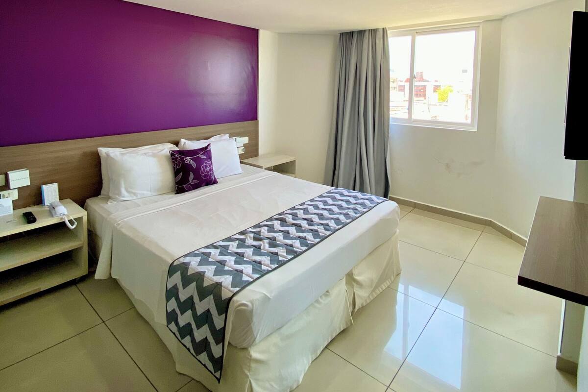 A hotel room with a bed, a nightstand, a TV, and a window with curtains.