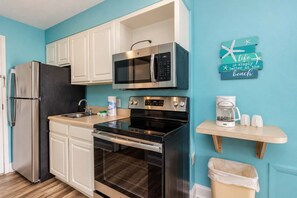 A compact kitchen with white cabinets, a stainless steel refrigerator, a microwave, an oven, and a coffee maker on a wooden shelf.