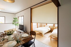 Une chambre d’hôtel moderne avec un grand lit, un coin repas avec une table dressée pour deux personnes, et une grande fenêtre offrant une vue agréable.