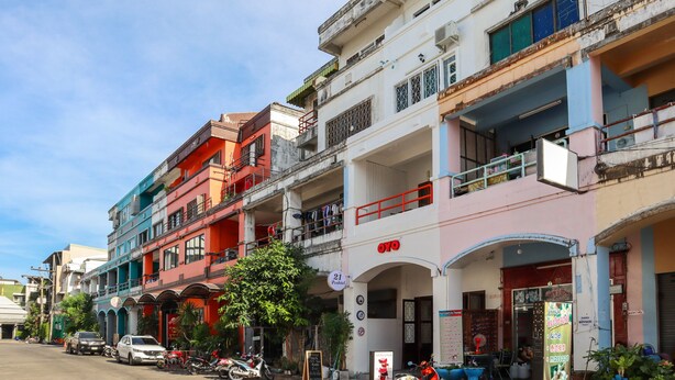 A row of colorful buildings with a sign for 'OYO' hotel. There are parked vehicles and a few people visible.