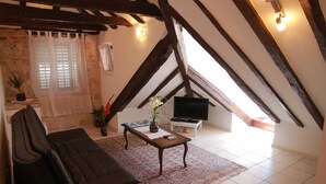 Una sala de estar acogedora con un sofá de cuero oscuro, una mesa de centro de madera y un televisor. Hay una ventana con cortina translúcida, una planta en maceta y un piso de baldosas.