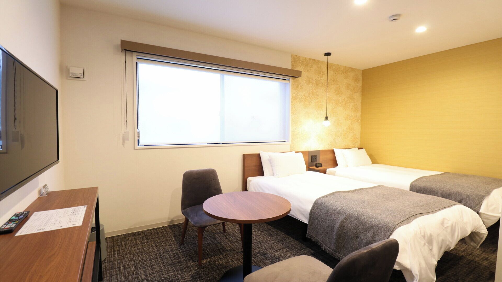 Quarto de hotel com duas camas, uma escrivaninha com cadeira, televisão e uma janela grande.