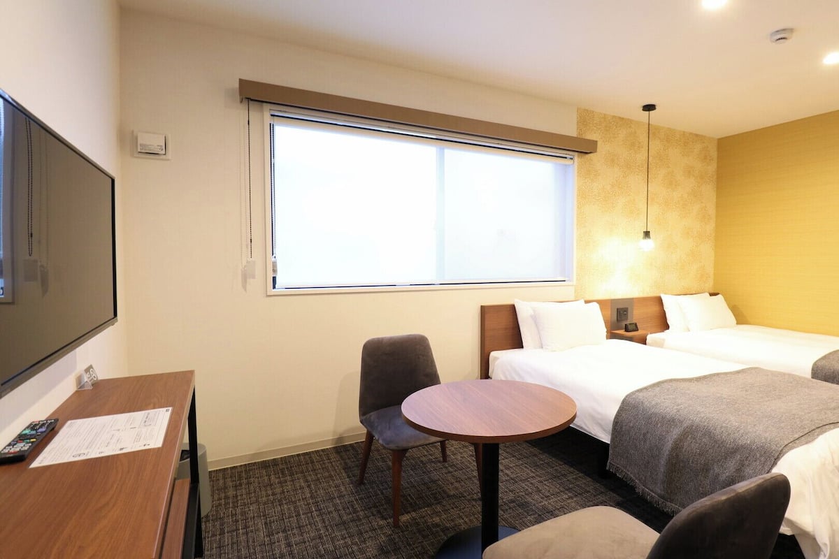 A hotel room with two beds, a desk with a chair, a TV, and a large window.