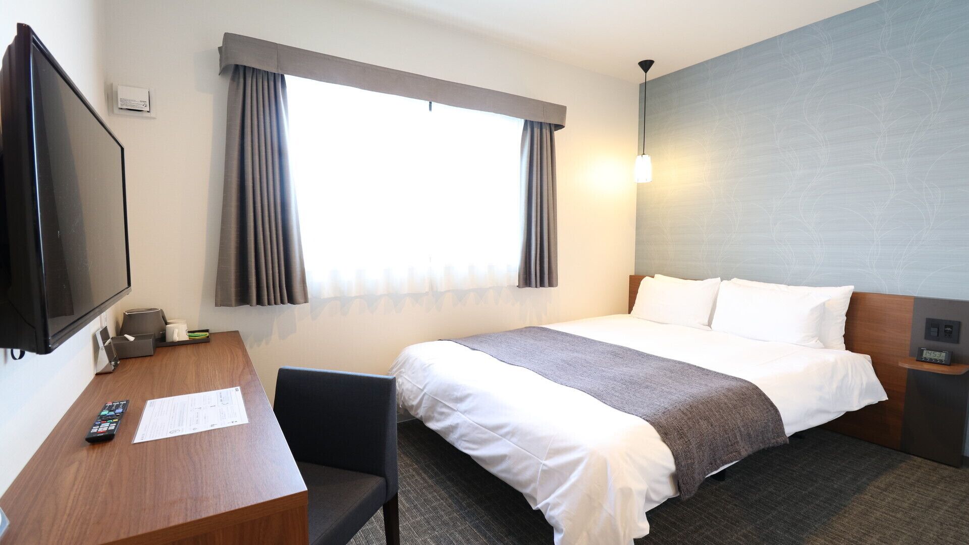 Quarto de hotel com cama, escrivaninha com televisão, cadeira, janela com cortinas e relógio de parede.