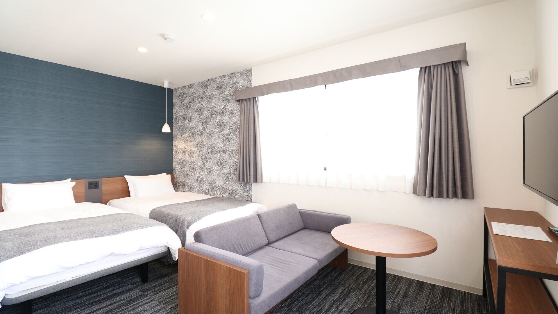 Quarto de hotel com duas camas, um sofá, uma mesinha, uma televisão e uma janela com cortinas.
