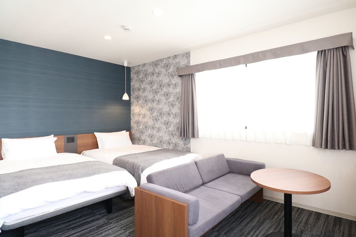 A hotel room with two beds, a sofa, a small table, a TV, and a window with curtains.