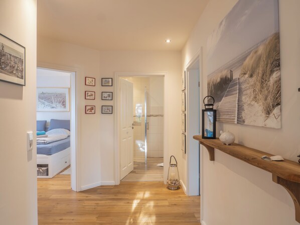A hallway with wooden flooring, a bed with a blue mattress, a door leading to a bathroom, and a wall with framed pictures and a lantern.