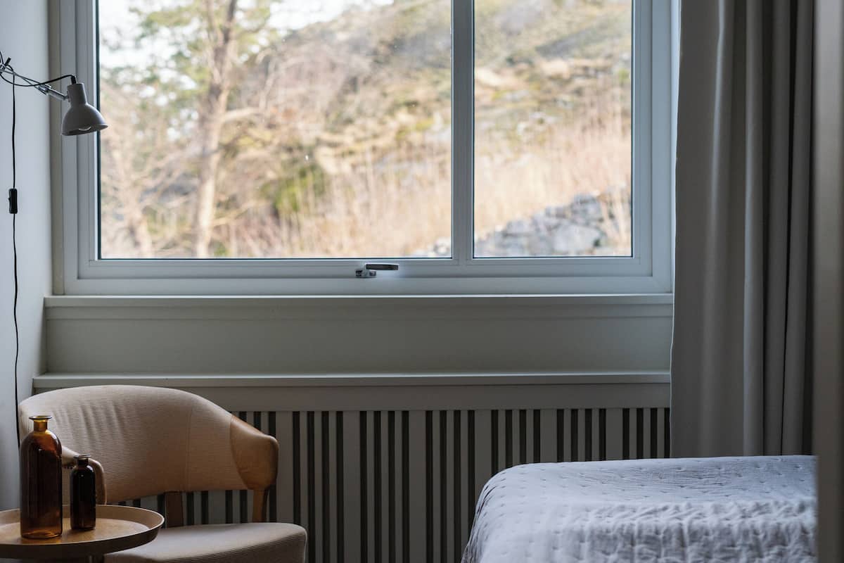 Un dormitorio con una ventana grande, una silla, una mesita de noche con botellas y una cama con cubierta blanca.