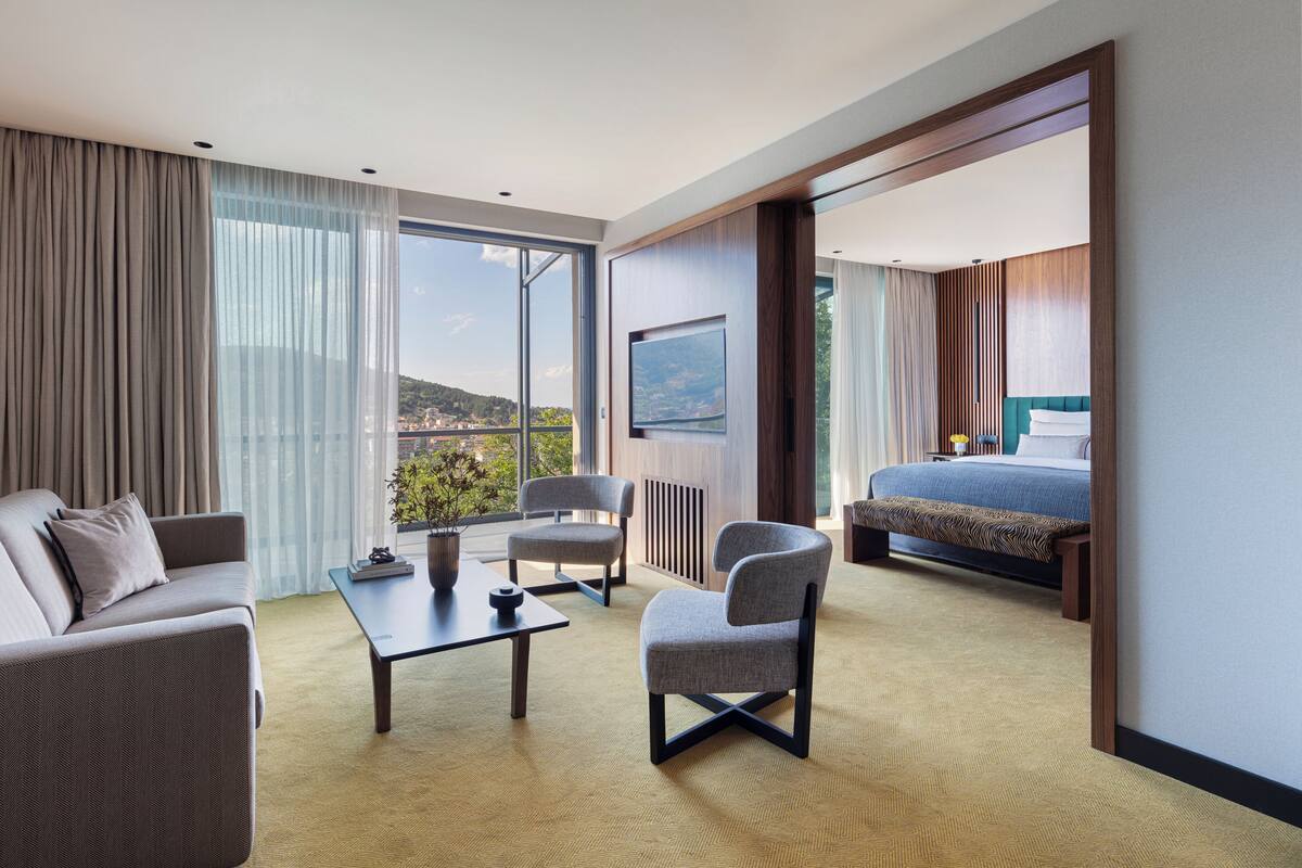 A modern hotel room with a sofa, armchairs, a coffee table, a bed, and a large window offering a city view.