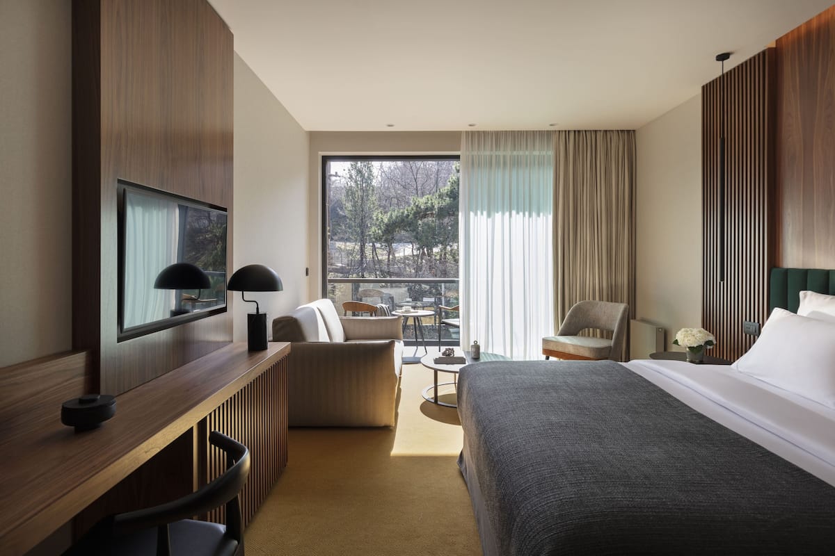 A hotel room with a bed, a desk with a computer, a chair, a small table, and a view of the outdoors.