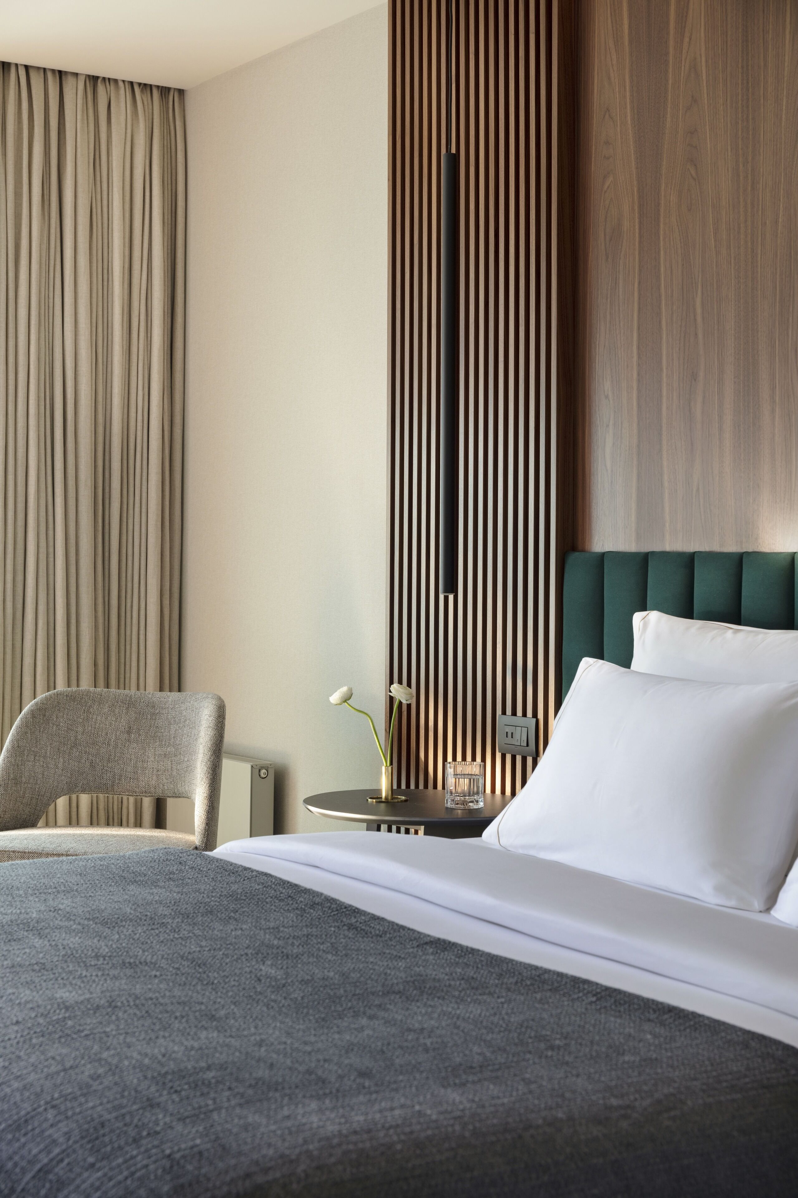 A hotel room with a bed, a chair, a small table with a vase, and a wall with vertical wooden slats.