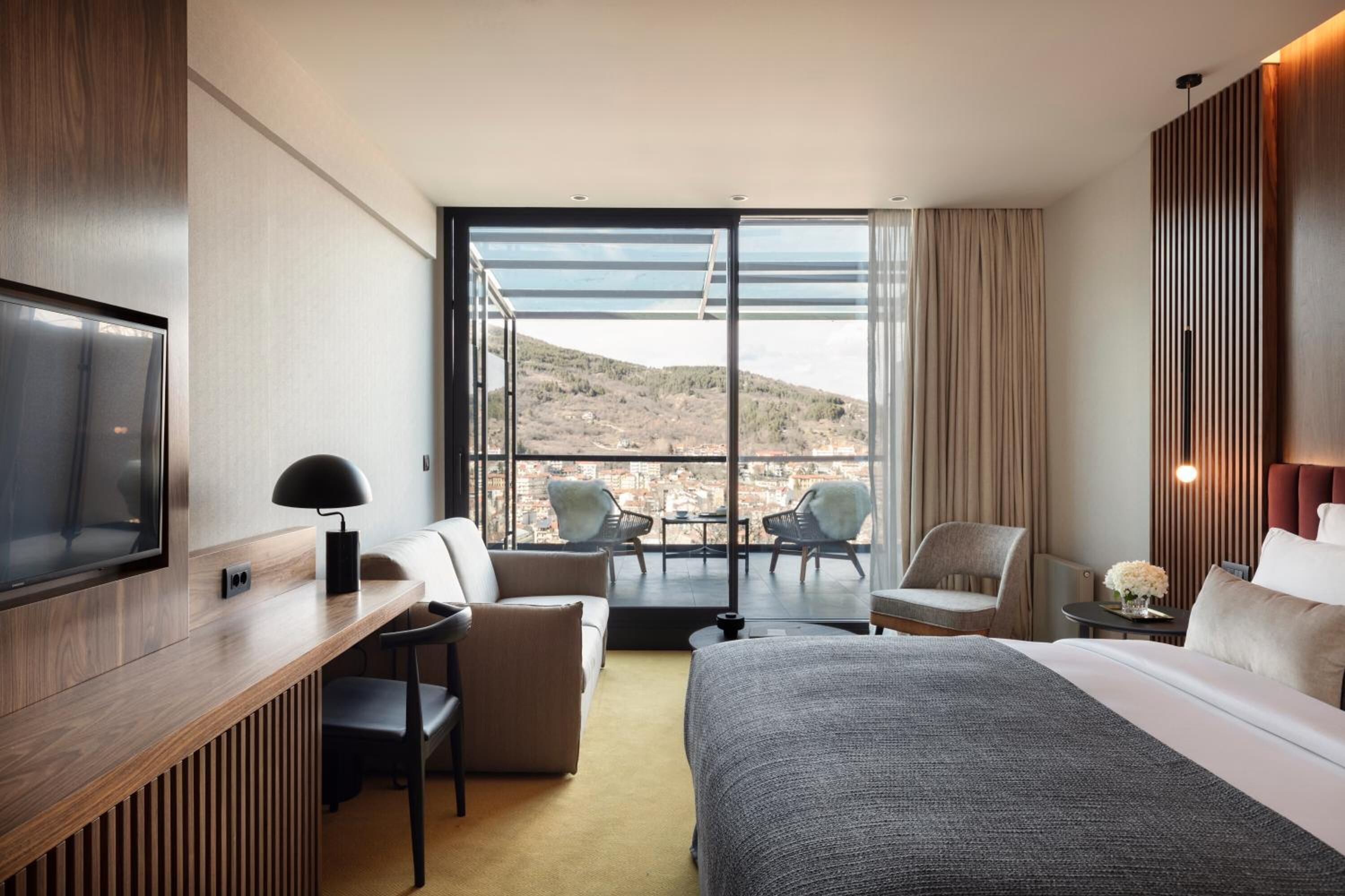 Ein modernes Hotelzimmer mit einem großen Bett, einem Schreibtisch, zwei Stühlen, einem Fernseher und einem Balkon mit Blick auf die Stadt und die Hügel.