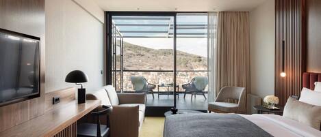 Habitación de hotel moderna con una cama grande, un escritorio, dos sillas, un televisor y un balcón con vistas a la ciudad y las colinas.