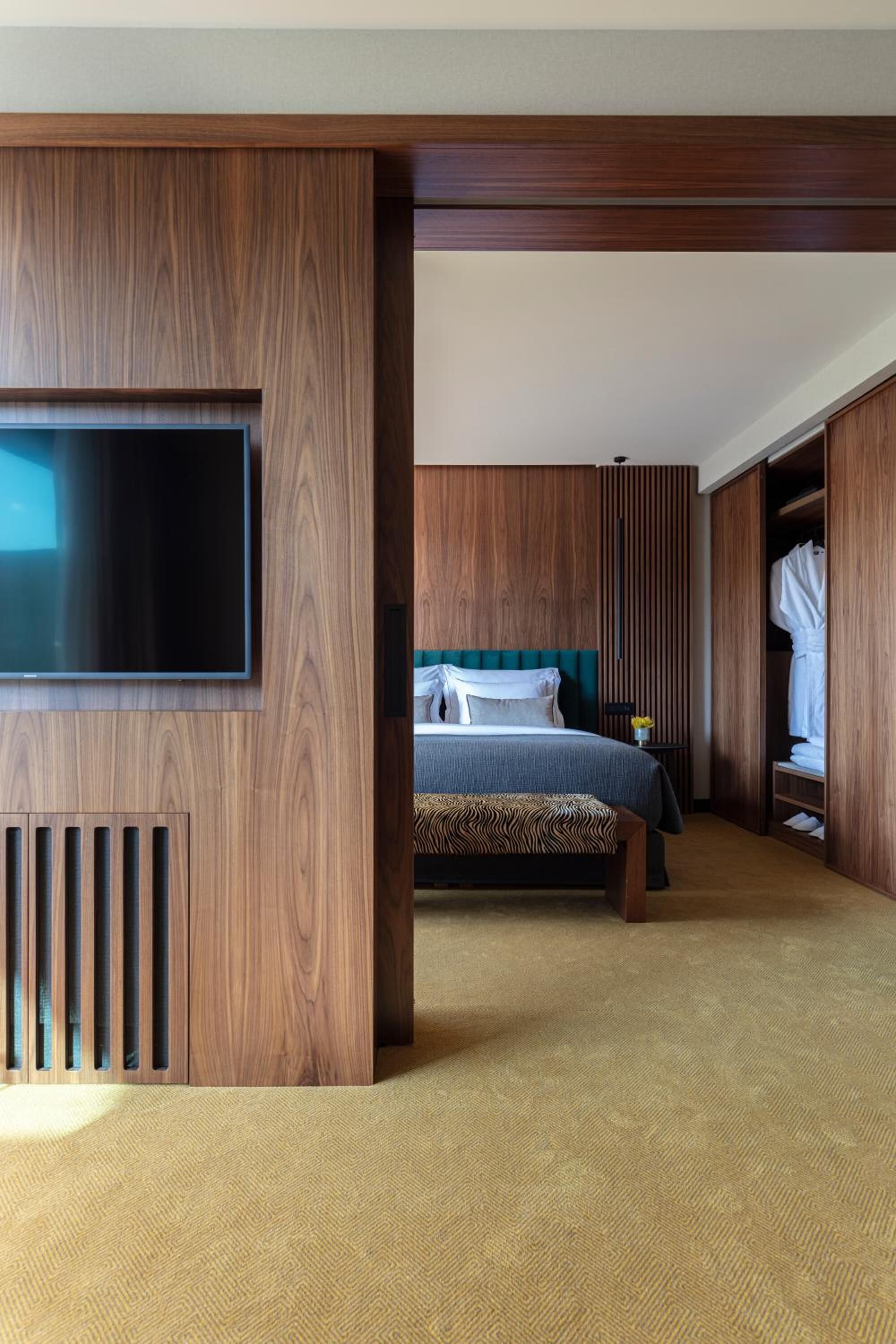 Habitación de hotel con una amplia televisión de pantalla plana montada en un panel de madera, una cama con cabecera verde, un armario de madera y una pequeña mesa con un florero.