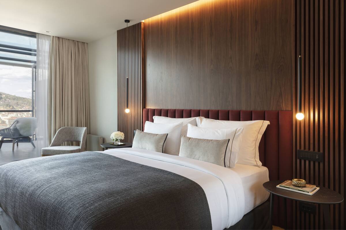 A modern hotel room with a large bed, a wooden accent wall, a nightstand, and a view of the outdoors.