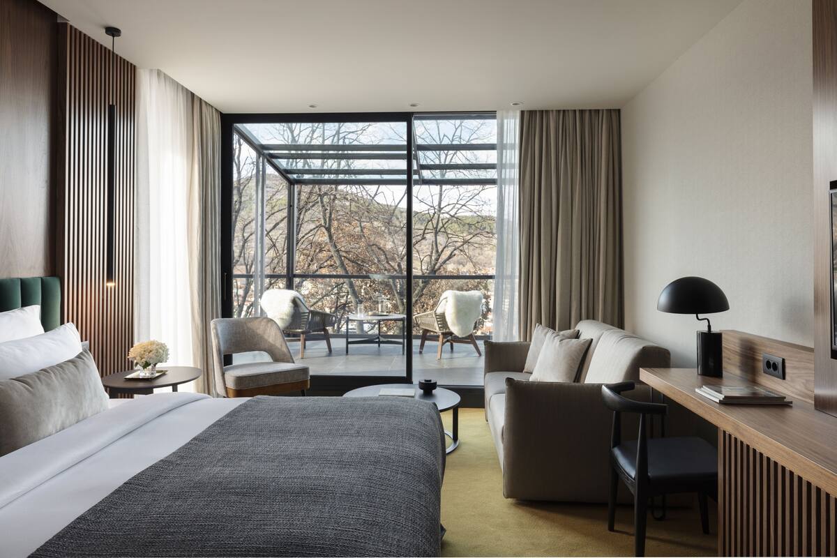 A modern hotel room with a large bed, a desk, a sofa, and a balcony with a view of trees and mountains.