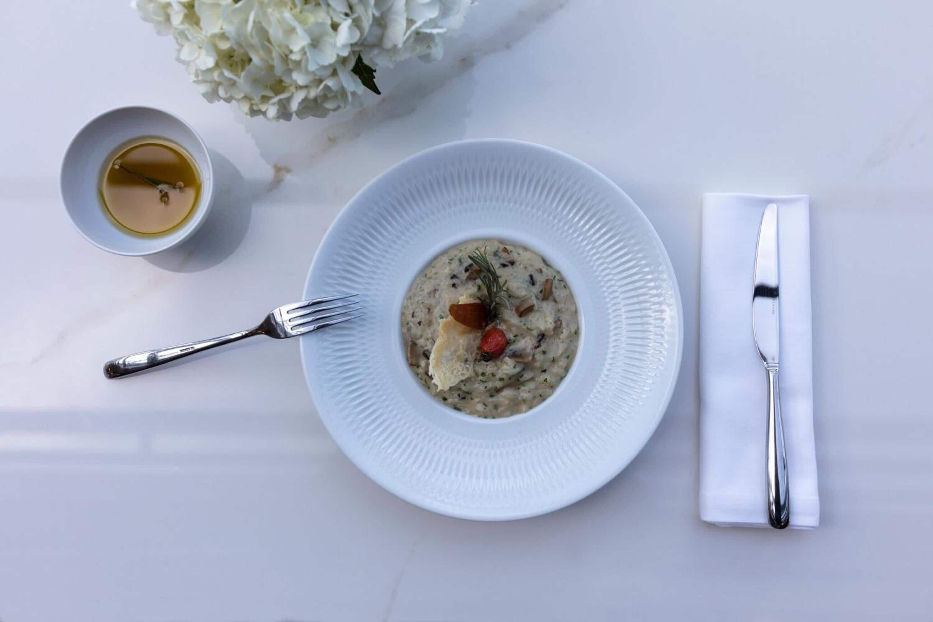 A white plate with a creamy soup garnished with herbs and a small piece of red fruit, accompanied by a fork and a napkin on a white table.