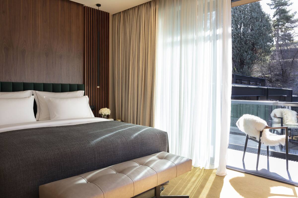 A modern bedroom with a large bed, a bench, a nightstand with a vase, and a view of a balcony with chairs.