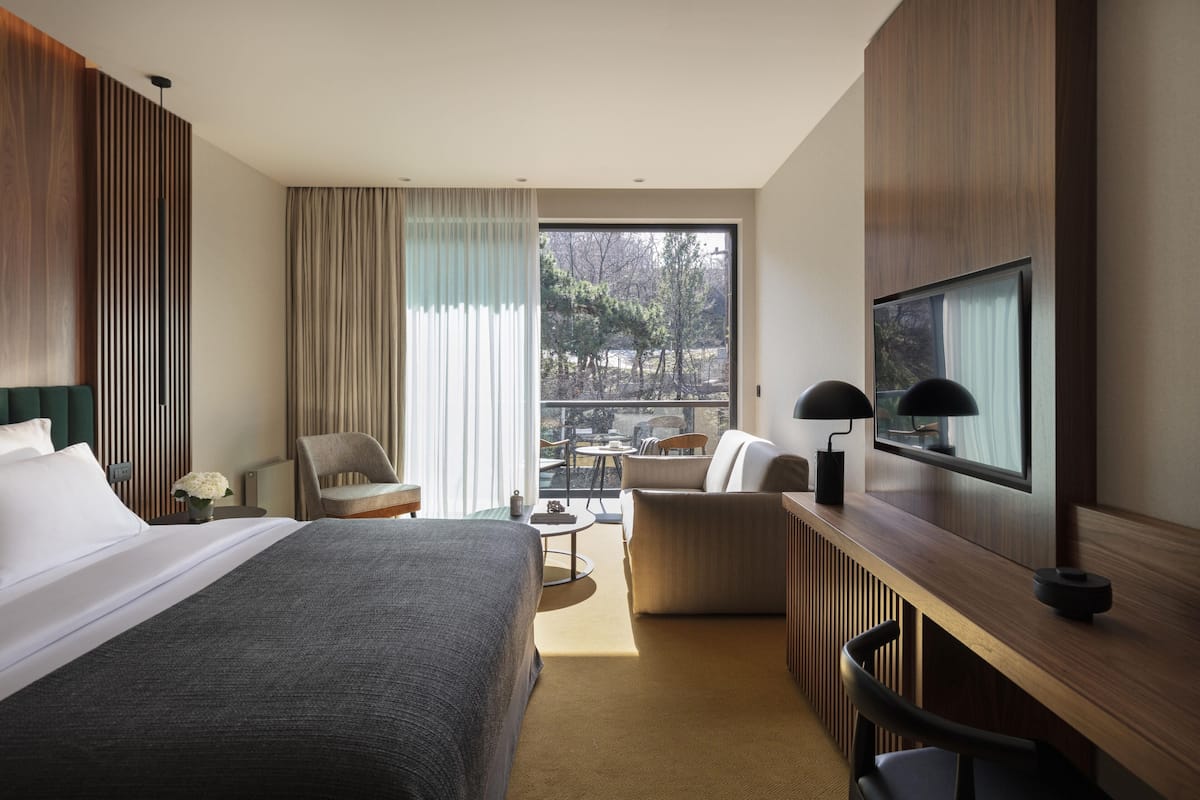 A hotel room with a large bed, a desk with a TV, a seating area with chairs, and a view of the outdoors through the window.