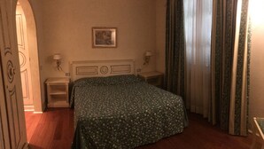 Une chambre d’hôtel avec un lit, deux tables de chevet, une petite table, un tableau encadré au mur et une fenêtre avec des rideaux.