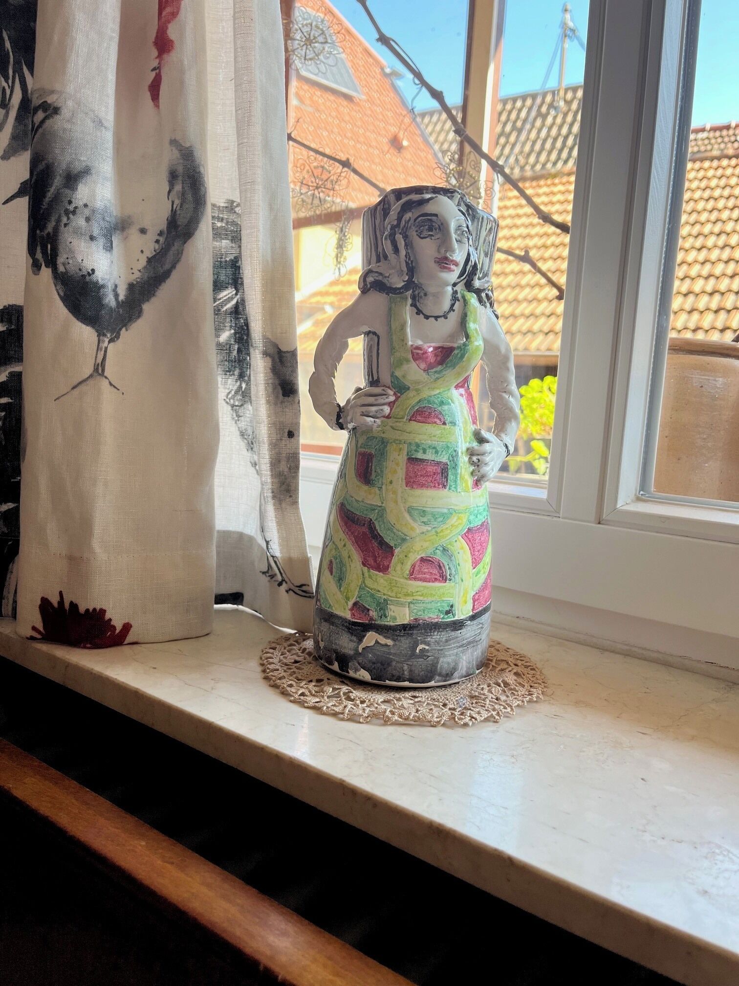 Une figurine posée sur un rebord de fenêtre, d’où l’on aperçoit une maison et un arbre à l’extérieur.